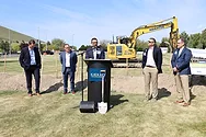 People speaking at podium in a grassy field with a digger.