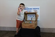 A kid standing next to a treasure chest with a sign that says, "Treasure Trove".