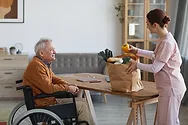A nurse with a grocery bag and a man in a wheelchair.
