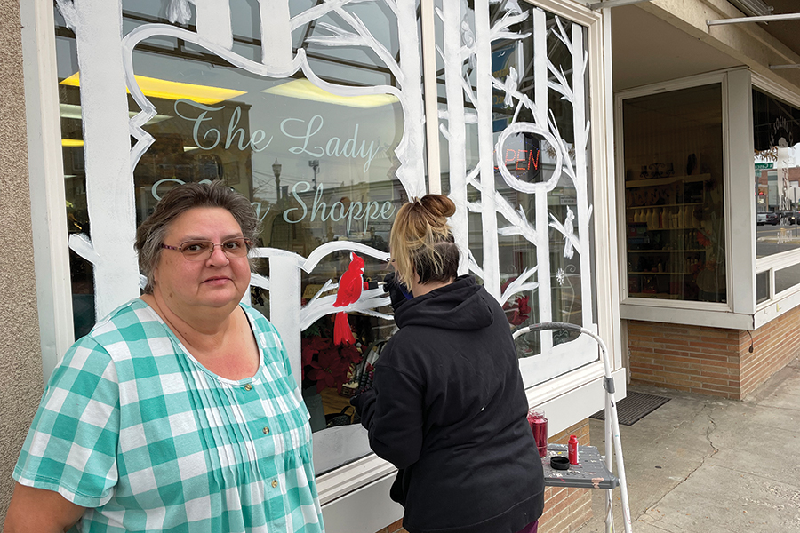 Cindy Mosley-Cleary, owner of The Lady Bug Shoppe in downtown Kennewick, poses in front of her store at 304 W. Kennewick Ave., as Alicia Michaliszyn of Allusions Art & Design paints a winter scene featuring glittery red cardinals, a symbol of hope — and Mosley-Cleary’s hopes for a successful shopping season. (Photo by Kristina Lord)