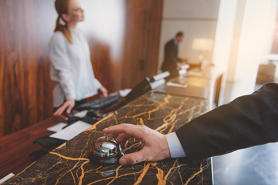 Male hand ringing hotel bell at counter with index finger. Female receptionist standing on background